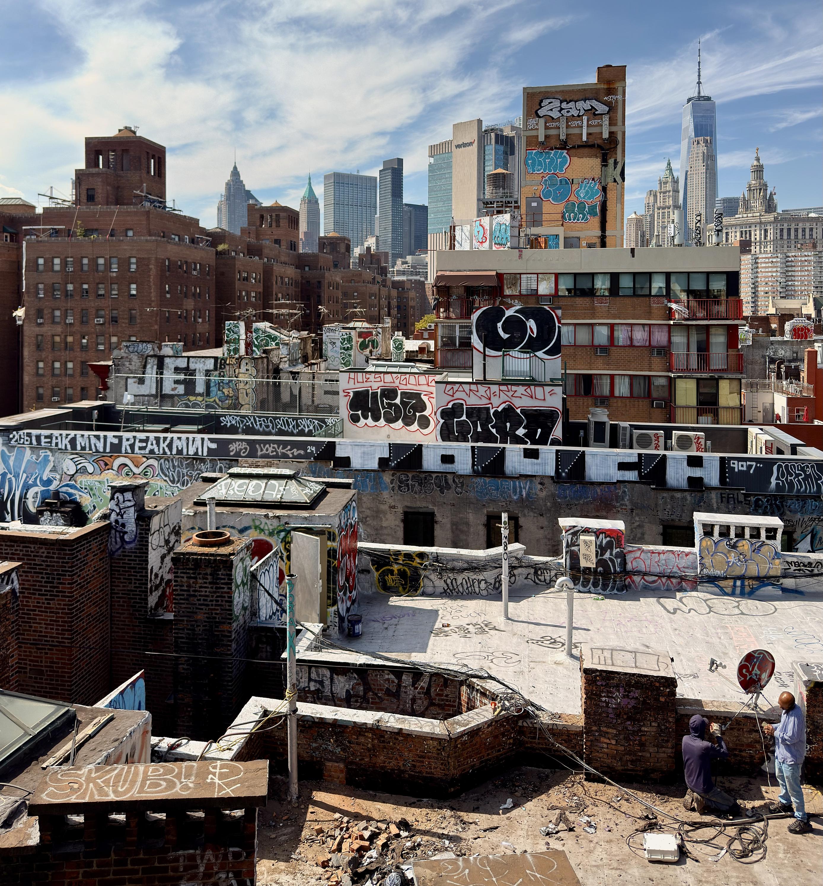 A color photograph of a rooftop view in the Two Bridges neighborhood of lower Manhattan on a bright day. The foreground shows many cluttered roofs covered in rubble and various vent pipes and brick enclosures. The walls and many of the rooftop structures are heavily covered in graffiti tags. In the bottom right corner, two individuals are working. One person, wearing a hoodie and shorts, kneels on the rooftop, while another person stands beside them. Wires and equipment are scattered on the ground around them. The skyline is filled with a variety of skyscrapers under a partly cloudy sky, most notably One World Trade Center which towers above the rest.