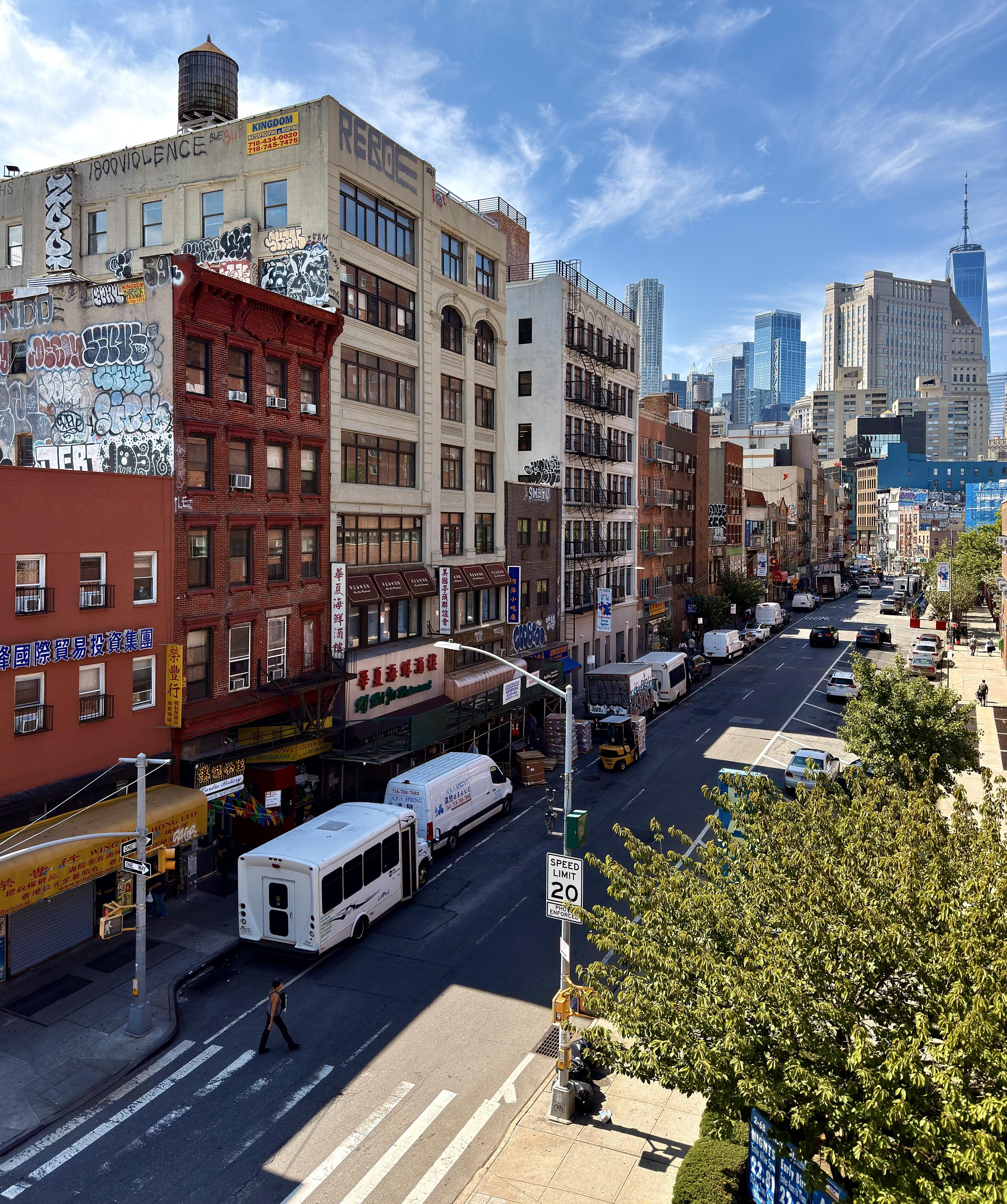 A color photograph of the street scene in Chinatown on a sunny day. The street, lined with multi-story brick and stucco buildings, stretches into the distance, with a dense mix of cars, trucks, and a small white bus visible. The sky is a bright blue with scattered clouds. On the left, buildings are covered in a variety of graffiti and street art, and some storefronts have colorful awnings. Atop the closest building on the left is a historic water tower. Signage in Chinese language lines both sides of the street. In the background, modern skyscrapers, including One World Trade Center, rise above the older buildings, contrasting with the foreground. The street is busy with activity, including cars driving, a pedestrian crossing a crosswalk in the lower left, and warehouse workers loading produce onto a fork lift. On the right side of the frame large green trees line the sidewalk.