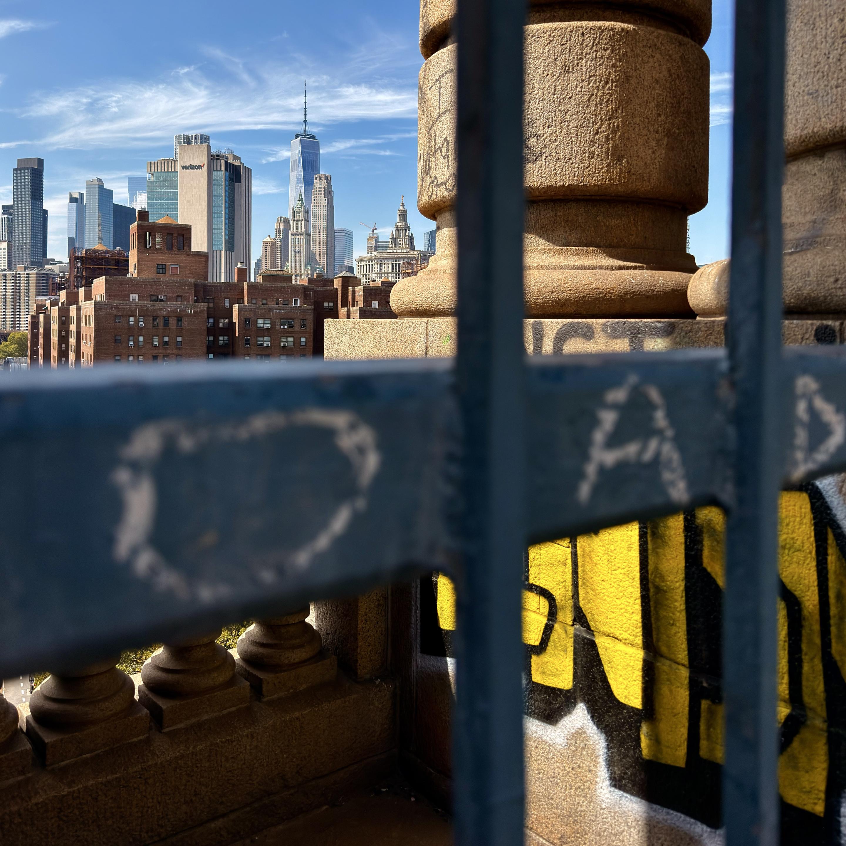 A color photograph in a square aspect ratio. It is an eye-level shot taken on a sunny afternoon from the Manhattan Bridge looking west toward the Financial District. The background features a detailed view of the lower Manhattan skyline under a clear blue sky with sparse white clouds. The One World Trade Center tower stands prominently in the center, flanked by other tall buildings, including the Verizon Building. In the foreground, a heavily weathered, dark-blue metal railing runs horizontally across the bottom and right side of the frame, partially obscuring the view. Below the railing, large, blocky yellow and black graffiti is spray-painted on a concrete base, with more subtle black graffiti visible on the concrete pier to the right. The large, rounded, stone columns of the bridge are visible on both the left and right sides of the photo, and small gaps between the columns reveal the tops of green trees on the ground below, adding a sense of depth.