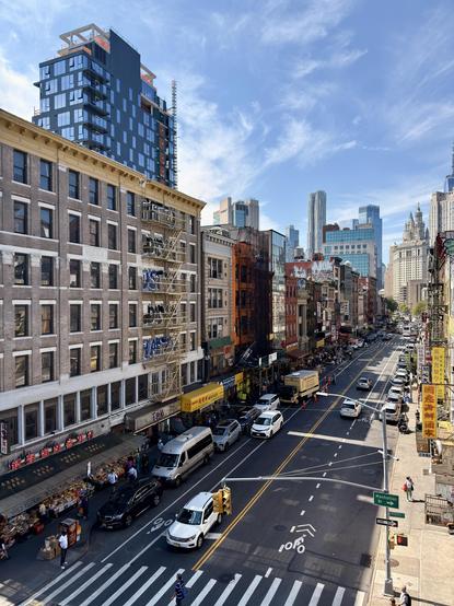 A color photograph in portrait orientation, a slightly elevated view of a bustling street in Chinatown, New York City, as seen from the pedestrian walkway on the Manhattan Bridge. The perspective looks down the street, which is lined on both sides with multi-story, classic brick and stone buildings, many of which have fire escapes, and graffiti on their facades. On the left side of the street, the buildings have a more traditional, older look, with street-level shops and Chinese language awnings line the street. Two of the storefronts are street markets with lots of produce displayed on tables outside and many people gathered around shopping. A tall, modern, dark blue high-rise building that is currently under construction stands prominently in the background behind the older structures on the left. The street itself is filled with a mix of vehicles, including cars, vans, and delivery trucks. The road has clear white lane markings and a prominent crosswalk in the foreground. In the distance, the iconic skyline of lower Manhattan is visible, including the Woolworth Building and One World Trade Center. The sky is bright blue with scattered white clouds.