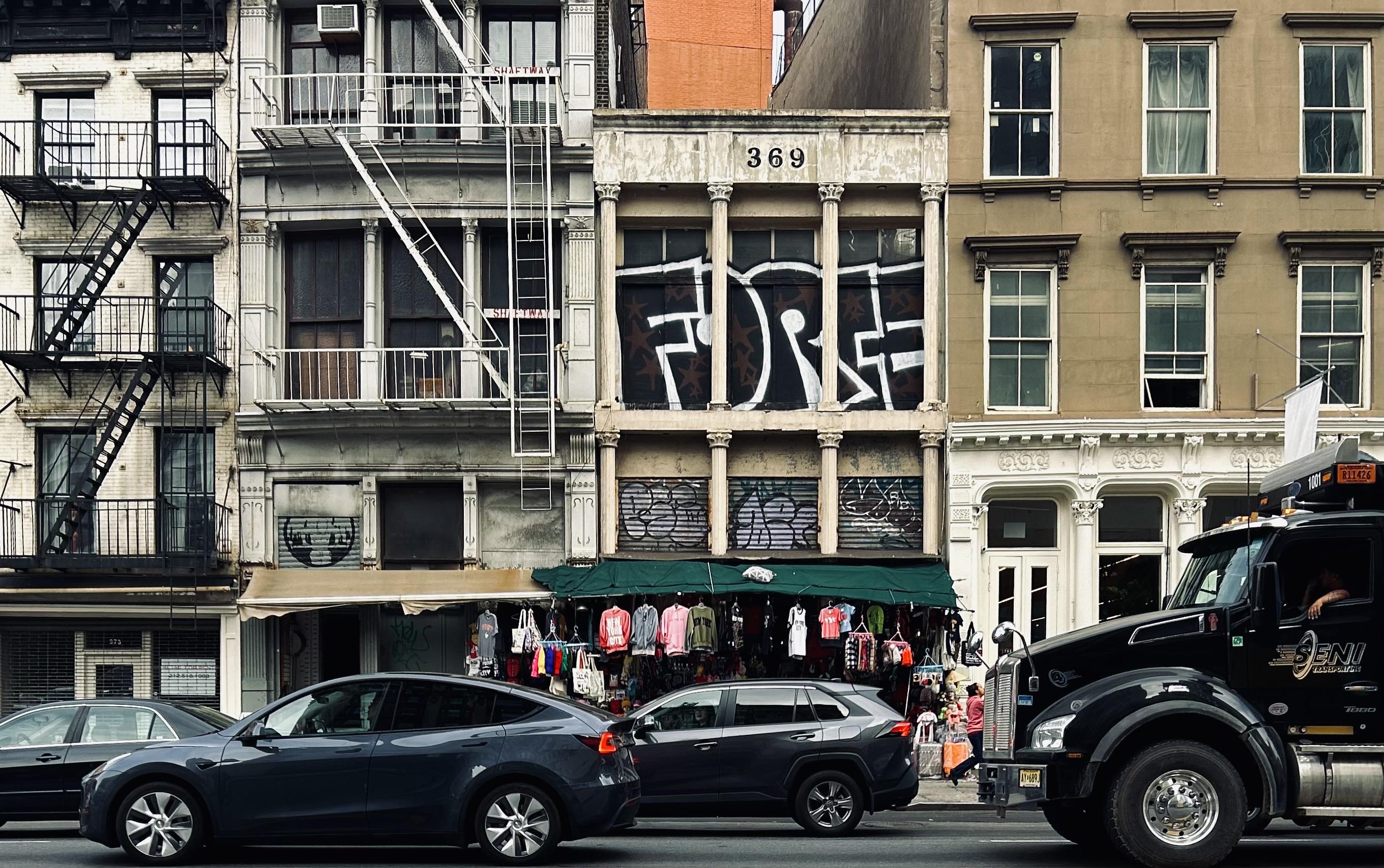A color photograph in landscape orientation, a street-level shot of the scene along Canal Street in the SoHo neighborhood of Manhattan on an overcast afternoon. The focal point of the image is a row of diverse buildings. In the foreground, cars drive by from left to right, including a large black semi-truck on the right. The driver of the semi-truck has their left arm hanging partially outside the driver side window. The buildings are a mix of architectural styles and heights. The most prominent one is in the center, a beige building with a "369" address marker and a large, black-and-white graffiti tag that reads "FORCE" across the second-story windows. Fire escapes are visible on the buildings to the left, and a shop with a green awning and clothes hanging outside is at the ground level of the central buildings.