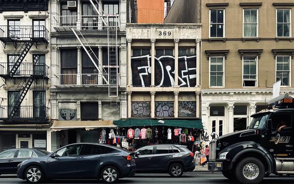 A color photograph in landscape orientation, a street-level shot of the scene along Canal Street in the SoHo neighborhood of Manhattan on an overcast afternoon. The focal point of the image is a row of diverse buildings. In the foreground, cars drive by from left to right, including a large black semi-truck on the right. The driver of the semi-truck has their left arm hanging partially outside the driver side window. The buildings are a mix of architectural styles and heights. The most prominent one is in the center, a beige building with a "369" address marker and a large, black-and-white graffiti tag that reads "FORCE" across the second-story windows. Fire escapes are visible on the buildings to the left, and a shop with a green awning and clothes hanging outside is at the ground level of the central buildings.