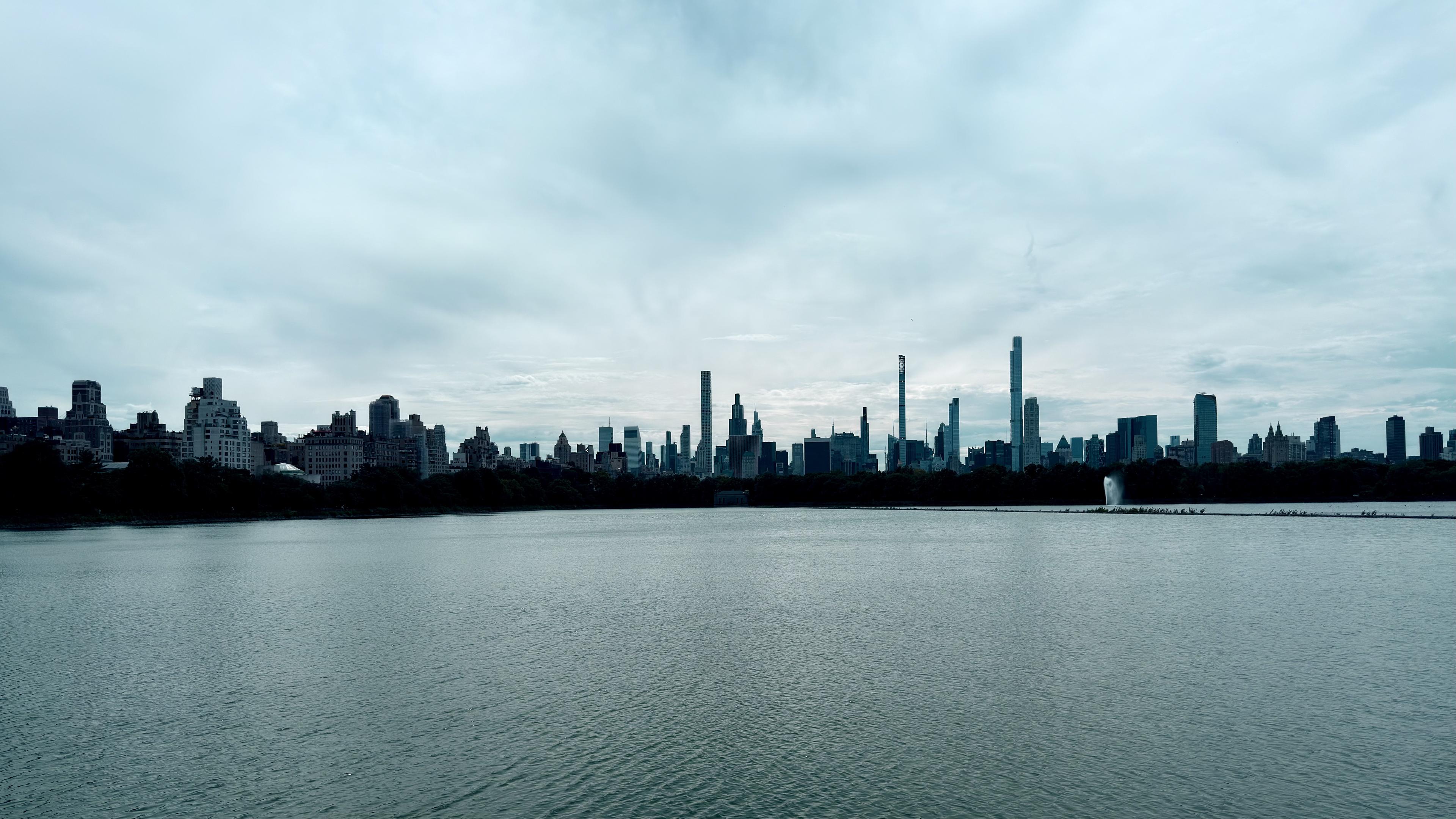 A panoramic, wide-angle shot of the Jacqueline Kennedy Onassis Reservoir in Central Park, New York City, taken from the 97th Street transverse side. The calm, dark blue-grey water fills the foreground and mid-ground. The surface of the water is slightly rippled under a vast, cloud-filled sky with soft, diffused light. The skyline of Manhattan is visible on the far side of the reservoir, forming a distinct silhouette of residential buildings and skyscrapers against the horizon. It has recently been raining, and the image has a cool, desaturated color palette, dominated by shades of blue, grey, and teal.