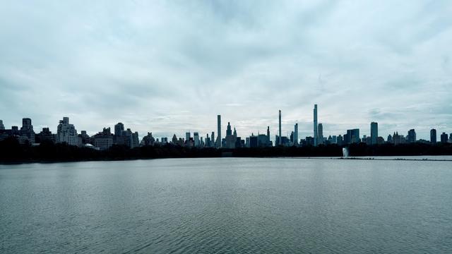 A panoramic, wide-angle shot of the Jacqueline Kennedy Onassis Reservoir in Central Park, New York City, taken from the 97th Street transverse side. The calm, dark blue-grey water fills the foreground and mid-ground. The surface of the water is slightly rippled under a vast, cloud-filled sky with soft, diffused light. The skyline of Manhattan is visible on the far side of the reservoir, forming a distinct silhouette of residential buildings and skyscrapers against the horizon. It has recently been raining, and the image has a cool, desaturated color palette, dominated by shades of blue, grey, and teal.