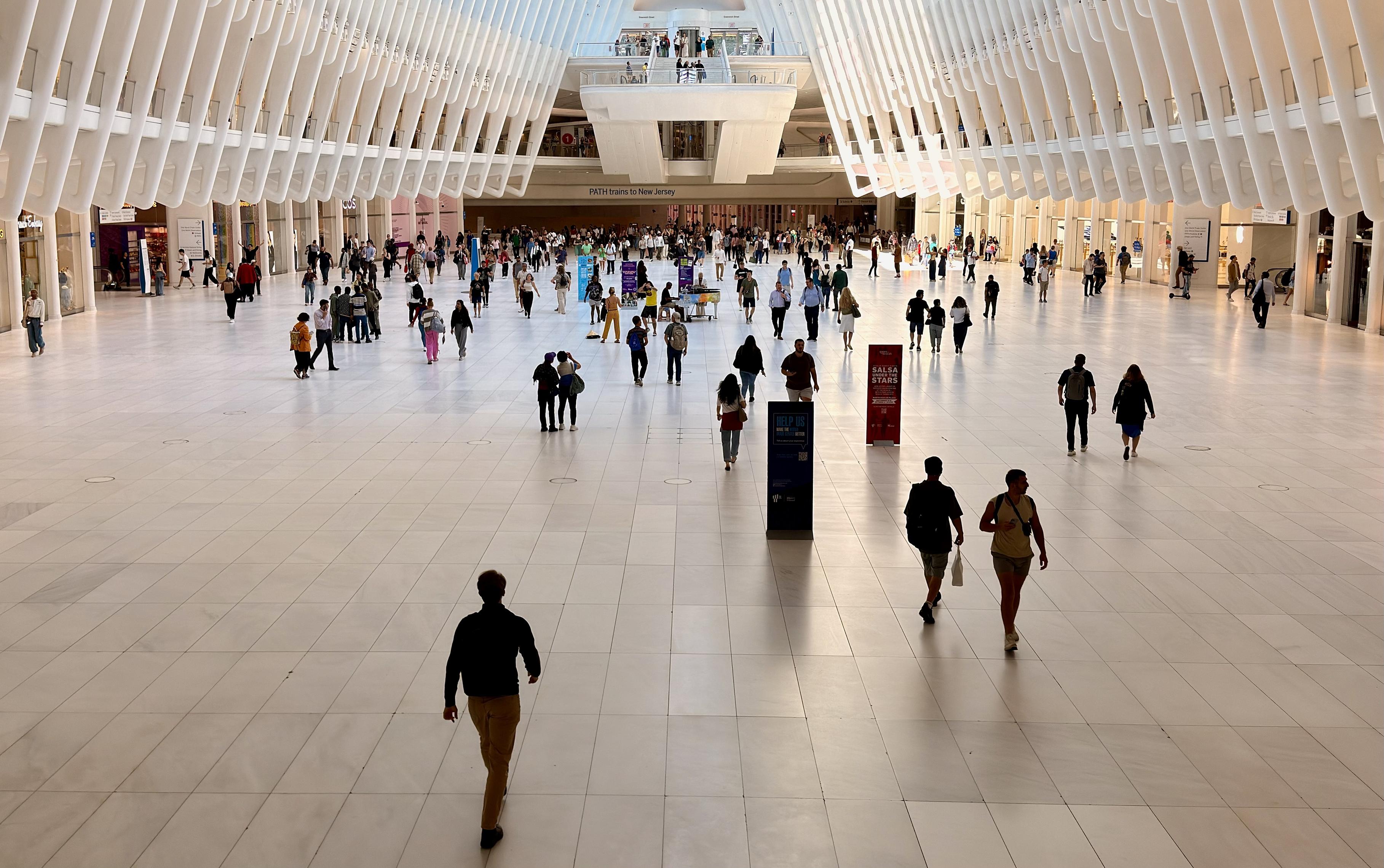 A color photograph in landscape orientation. An eye-level, wide shot of the lower level main hall of the Oculus at the World Trade Center in New York City. The space is bright and expansive, with a white, ribbed ceiling that resembles a skeletal structure. Tall, white ribs extend from the ceiling down to the floor, creating a sense of height and allow morning light to fill the space. The floor is a light gray and white tile. Numerous people are walking in different directions across the floor, some individually and others in small groups. In the center of the image, an observation deck extends over the main hall and many people are walking by and standing taking photos . In the background, there are a few kiosks and the entrance to the PATH trains, with a sign that reads "PATH Trains to New Jersey".