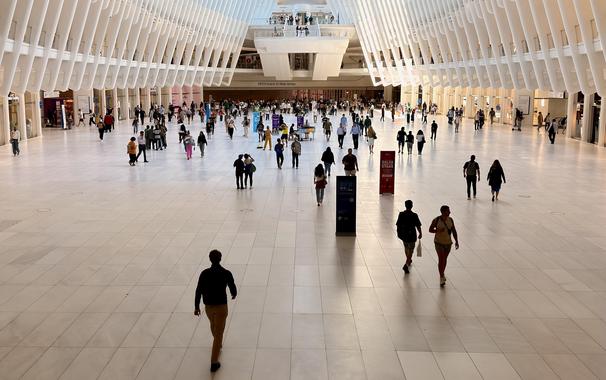 A color photograph in landscape orientation. An eye-level, wide shot of the lower level main hall of the Oculus at the World Trade Center in New York City. The space is bright and expansive, with a white, ribbed ceiling that resembles a skeletal structure. Tall, white ribs extend from the ceiling down to the floor, creating a sense of height and allow morning light to fill the space. The floor is a light gray and white tile. Numerous people are walking in different directions across the floor, some individually and others in small groups. In the center of the image, an observation deck extends over the main hall and many people are walking by and standing taking photos . In the background, there are a few kiosks and the entrance to the PATH trains, with a sign that reads "PATH Trains to New Jersey".