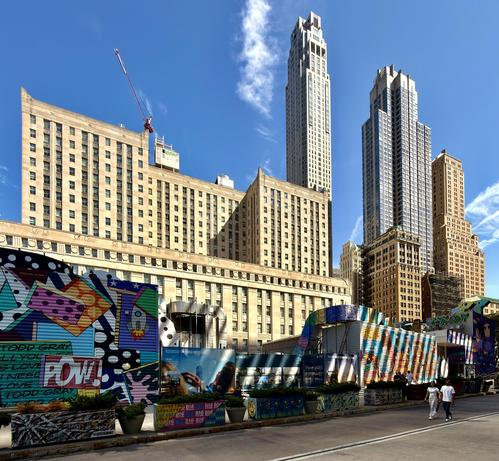 A color photograph of the street scene outside of The Oculus, taken on a clear, sunny day. The foreground features a long, colorful barricade covered in various artistic styles, including graffiti-like patterns, abstract shapes, and posters. Two pedestrians are walking along the sidewalk to the right, in front of the barricade. One of the people is looking up at the sky towards One World Trade Center. Behind the barrier is a large, classic building with a light-colored stone facade and many windows, and a red construction crane is visible on a roof behind it. In the background, a cluster of modern skyscrapers with reflective glass and unique architectural designs towers over the street. The sky is bright blue with scattered white clouds, and the sun casts sharp shadows on the buildings.