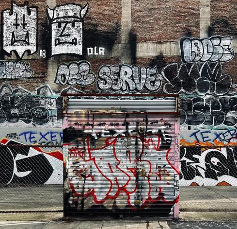 A color photograph in a square aspect ratio of a vacant parking lot on the corner of Lafayette Street at Canal Street in the SoHo neighborhood of Manhattan. It is afternoon time on a partly cloudy day. The image shows a detailed view of a brick wall, and in front of it a metal garage door, both covered in a dense mix of graffiti. The upper part of the brick wall features a series of graffiti pieces. The scene is framed by a chain-link fence in the foreground on either side of the garage door. The lower part of the image is dominated by a metal garage door, which is covered in a chaotic combination of tags. The scene is still.