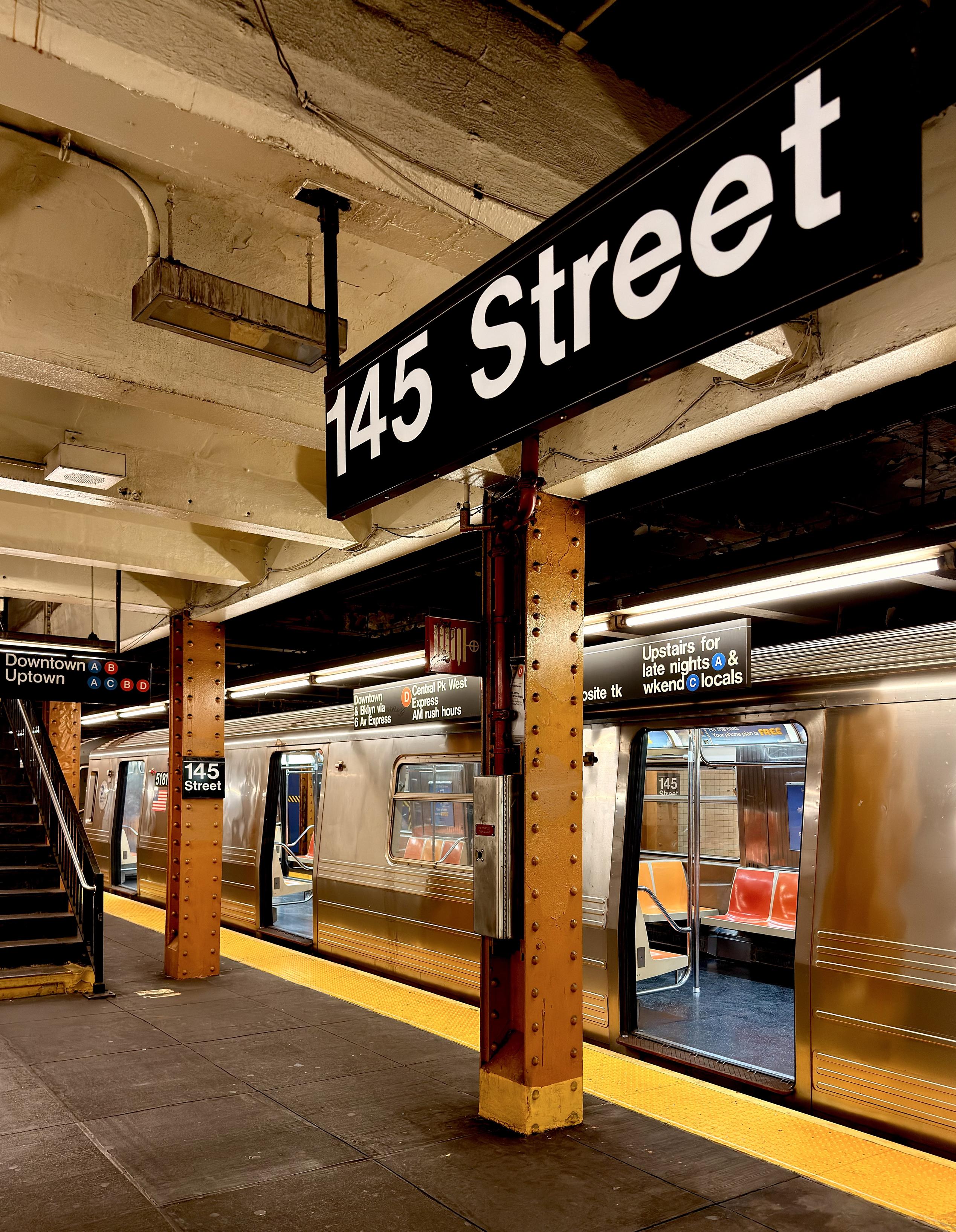 A color photograph taken from a low angle on a subway platform at the 145th Street station in Manhattan. The foreground is dominated by a large, black station sign with the white text "145 Street" mounted to a support pillar. The pillar is a textured, brown metal with visible rivets. A bright yellow safety strip runs along the edge of the platform. A train with open doors is stopped at the platform on the right side of the image. Its metallic exterior reflects the warm, yellowish light from the station's overhead lights. On the left, a set of stairs leads up to a different part of the station. A smaller sign above the stairs reads "Downtown Uptown” and there are train line iconography. The ceiling above the platform is a mix of exposed pipes, lights, and a faded, yellowish-brown surface, giving the scene a warm, vintage feel.