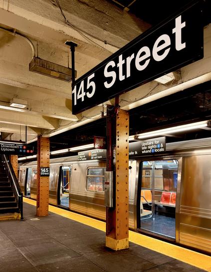 A color photograph taken from a low angle on a subway platform at the 145th Street station in Manhattan. The foreground is dominated by a large, black station sign with the white text "145 Street" mounted to a support pillar. The pillar is a textured, brown metal with visible rivets. A bright yellow safety strip runs along the edge of the platform. A train with open doors is stopped at the platform on the right side of the image. Its metallic exterior reflects the warm, yellowish light from the station's overhead lights. On the left, a set of stairs leads up to a different part of the station. A smaller sign above the stairs reads "Downtown Uptown” and there are train line iconography. The ceiling above the platform is a mix of exposed pipes, lights, and a faded, yellowish-brown surface, giving the scene a warm, vintage feel.