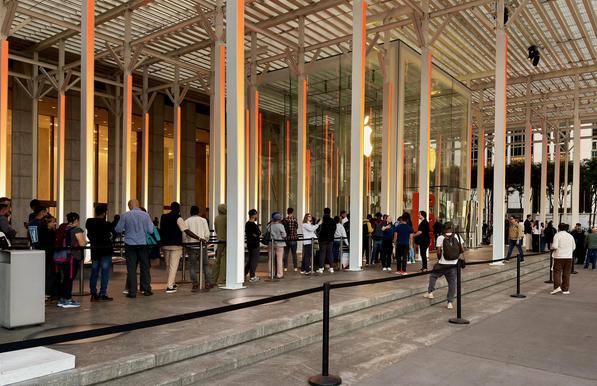 A color photograph in landscape aspect ratio. The shot captures a crowd of people lined up outside the Apple Store on Fifth Avenue in New York City early in the morning. The store's entrance, a striking 32-foot transparent glass cube, is the central focus of the image, with a prominent Apple logo visible inside. The store is located on a public plaza, surrounded by a metal and glass canopy structure supported by tall, orange-lit columns to commemorate the launch of the iPhone 17. People are shown waiting in a queue behind a black rope barrier waiting for the store to open. The plaza features wide concrete steps and metal railings that guide the flow of the crowd. In the background, part of the General Motors Building is visible, providing the urban backdrop for the scene.