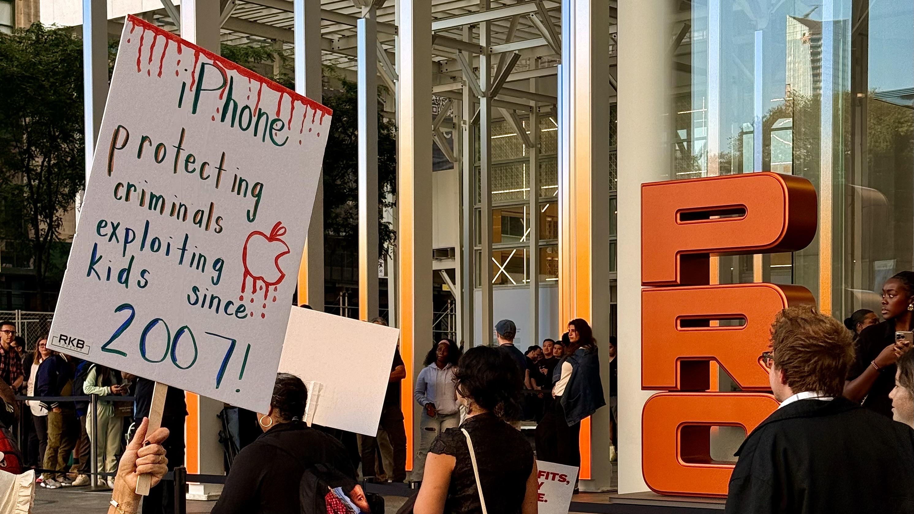 A color photograph taken of a protest outside the Fifth Avenue Apple store. Many people are marching in a circle. A person holds a white sign with a bloody effect at the top and the handwritten message: "iPhone protecting criminals exploiting kids since 2007!" with an Apple logo in the middle. In the background, there's a crowd of people, some holding smaller signs. The large orange letters "PRO" are visible as part of the iPhone 17 Pro marketing promotion.