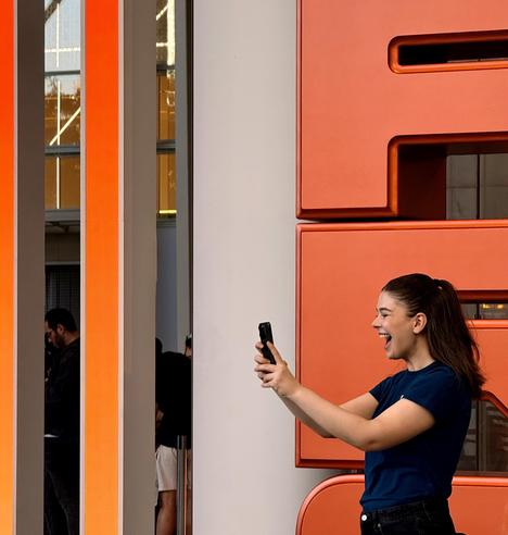 A cooor photograph of young woman, smiling widely with her mouth open in a laugh, holds a smartphone in both hands to take a selfie. She has long, dark brown hair tied in a high ponytail and is wearing a dark blue Apple retail employee t-shirt and black jeans. Her eyes are focused on the phone screen. In the background, there is a large, textured orange-red display to her right spelling out the word “PRO” and a reflective glass wall with vertical illuminated orange pillars to her left. A few people are partially visible in the background, out of focus. The scene is brightly lit by natural morning sunlight.