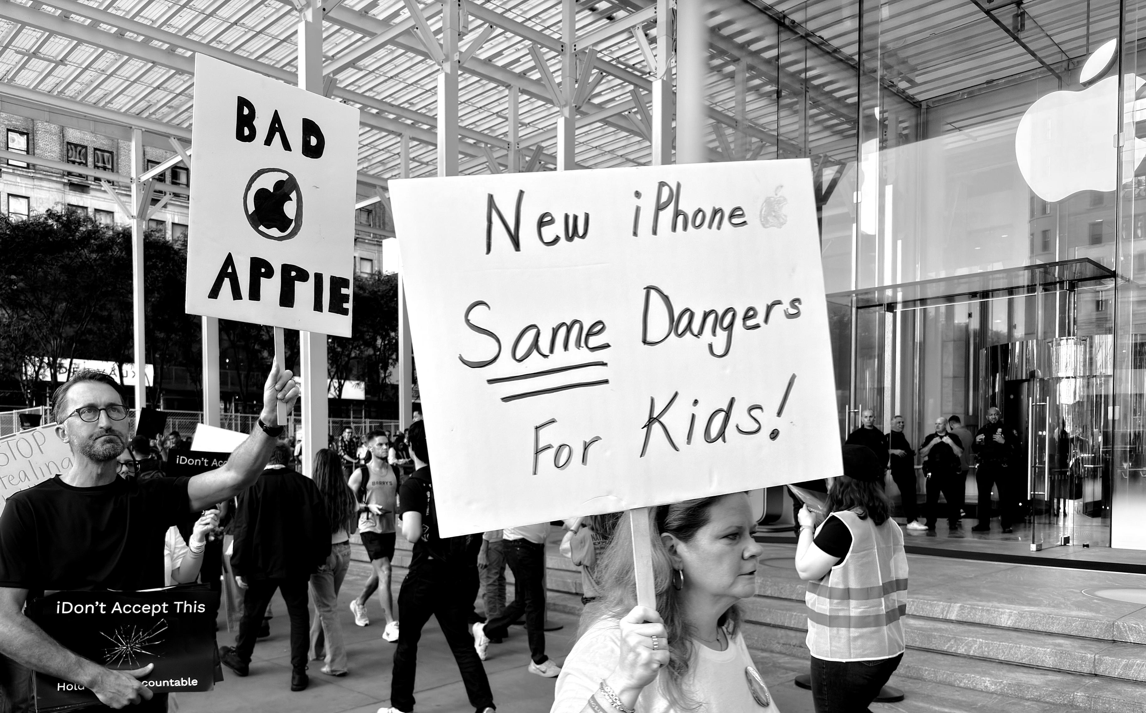 A high-contrast black and white photograph that captures a protest outside the Fifth Avenue Apple Store, identified by its large glass cube structure visible in the background. The protest is focused on two main figures holding large signs. The first sign has the words "BAD APPLE" written in bold, uppercase letters. A separate sign that reads "iDon't Accept This" and "Hold them accountable." The central figure is a woman, partially obscured by the main sign she is holding. The sign is large, white, and held horizontally. It has "New iPhone" with a small Apple logo, followed by a double-underlined "Same Dangers For Kids!" in large, hand-drawn letters. She is wearing a white collared shirt. Several other people are visible in the background, some holding signs or walking creating a crowd. The architecture of the Apple Store, with its glass and steel framework, provides the backdrop to the protest scene.