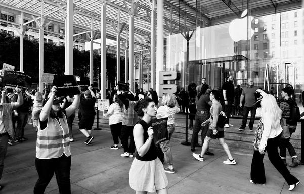 A black-and-white, wide-angle photograph taken at a protest outside the Apple Store on Fifth Avenue in New York City on a partly cloudy day. The image is taken from a low angle, showing the protest from a ground-level perspective. In the foreground, a woman wearing a light-colored, pleated skirt and a black, sleeveless top holds a dark protest sign. The sign is mostly obscured, but the word "Apple" is visible, followed by the text "Choose People Over Profits." Her head is turned to the left, and she looks contemplative. Behind her, a crowd of people are protesting, standing or walking along the sidewalk. The most prominent figure is a person in a safety vest and jeans, holding a protest sign with a partially obscured message that also mentions "Apple." Other individuals, some carrying signs, are visible further back. The background is dominated by the iconic glass cube entrance of the Apple Store, which is partially visible. The large, glowing Apple logo is visible on the glass facade, reflecting the cloudy sky. The rest of the scene is filled with the Fifth Avenue sidewalk, with other buildings and a few trees visible in the distance.