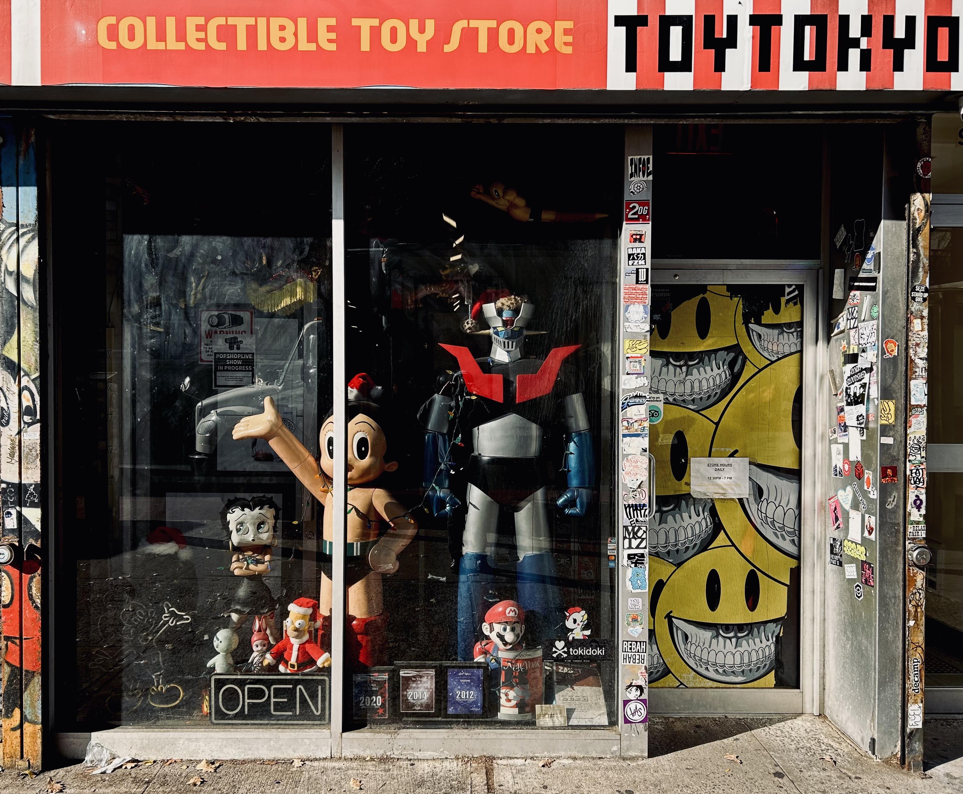A color photograph, eye-level street photo shows the storefront of Toy Tokyo, a collectible toy store in New York City's East Village. It is a bright sunny afternoon. The sign above the window reads "COLLECTIBLE TOY STORE" on a red banner with yellow block letters, and "TOYTOKYO" in black and red pixel-style lettering to the right of it. The large, grimy display window is crowded with giant character figures and smaller collectibles. The dominant figures are a towering robot with a red chest and blue legs, and a large, smiling, retro-styled Astro Boy with his right arm outstretched. Smaller figures are scattered on the floor. A small "OPEN" sign sits unlit on the floor at the bottom of the window. The window's glass reflects the street and sky, partially obscuring the view inside. To the right of the window is the entrance door, which is completely covered in stickers, posters, and graffiti. A large, stylized piece of street art is visible on the door, featuring a stack of several yellow, grinning smiley faces showing their teeth. Mickey Mouse has been added as graffiti to the wall to the left side of the store, but only a small vertical piece is visible. The dirty sidewalk creates the bottom edge of the image.