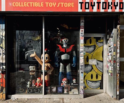 A color photograph, eye-level street photo shows the storefront of Toy Tokyo, a collectible toy store in New York City's East Village. It is a bright sunny afternoon. The sign above the window reads "COLLECTIBLE TOY STORE" on a red banner with yellow block letters, and "TOYTOKYO" in black and red pixel-style lettering to the right of it. The large, grimy display window is crowded with giant character figures and smaller collectibles. The dominant figures are a towering robot with a red chest and blue legs, and a large, smiling, retro-styled Astro Boy with his right arm outstretched. Smaller figures are scattered on the floor. A small "OPEN" sign sits unlit on the floor at the bottom of the window. The window's glass reflects the street and sky, partially obscuring the view inside. To the right of the window is the entrance door, which is completely covered in stickers, posters, and graffiti. A large, stylized piece of street art is visible on the door, featuring a stack of several yellow, grinning smiley faces showing their teeth. Mickey Mouse has been added as graffiti to the wall to the left side of the store, but only a small vertical piece is visible. The dirty sidewalk creates the bottom edge of the image.