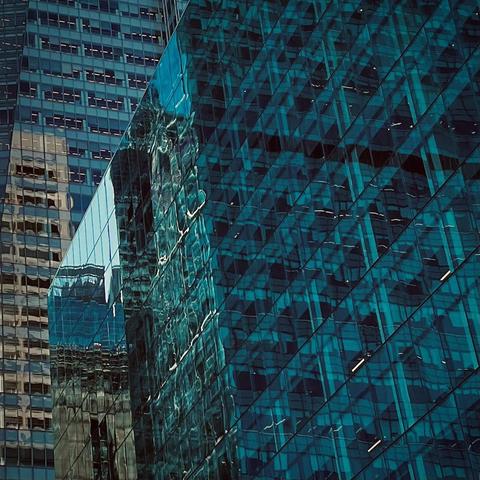 A color progressive of a highly detailed and textured close-up look at the modern, glass and steel exterior of skyscrapers in Times Square, Manhattan on a sunny afternoon. A building with a dark turquoise-blue glass facade, composed of a grid of reflective rectangular panels, dominates the right half of the frame. The glass has a wet, almost liquid appearance, with light streaking across its surface and creating a textured, almost abstract pattern. A reflection of the surrounding environment is visible, including the intricate grid of windows from an adjacent, lighter-colored building on the left.