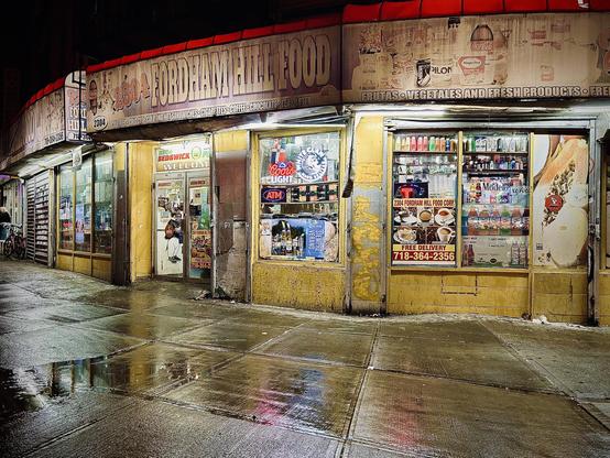 A color photograph of an exterior, eye-level shot of a convenience store called "Fordham Hill Food" at night. The storefront occupied a wide street corner lot. The store is brightly lit from within, casting a warm yellow glow onto the wet sidewalk and reflecting the light in the puddles. The storefront is a mix of glass windows and yellow siding. Various posters and advertisements cover the windows, including signs for an ATM, lottery, and free delivery. Inside, shelves are stocked with a wide range of drinks and other products. The top of the building has a red awning with white lettering that reads "Fordham Hill Food" and lists other products like cigarettes, fresh coffee, chocolate, and lotto.