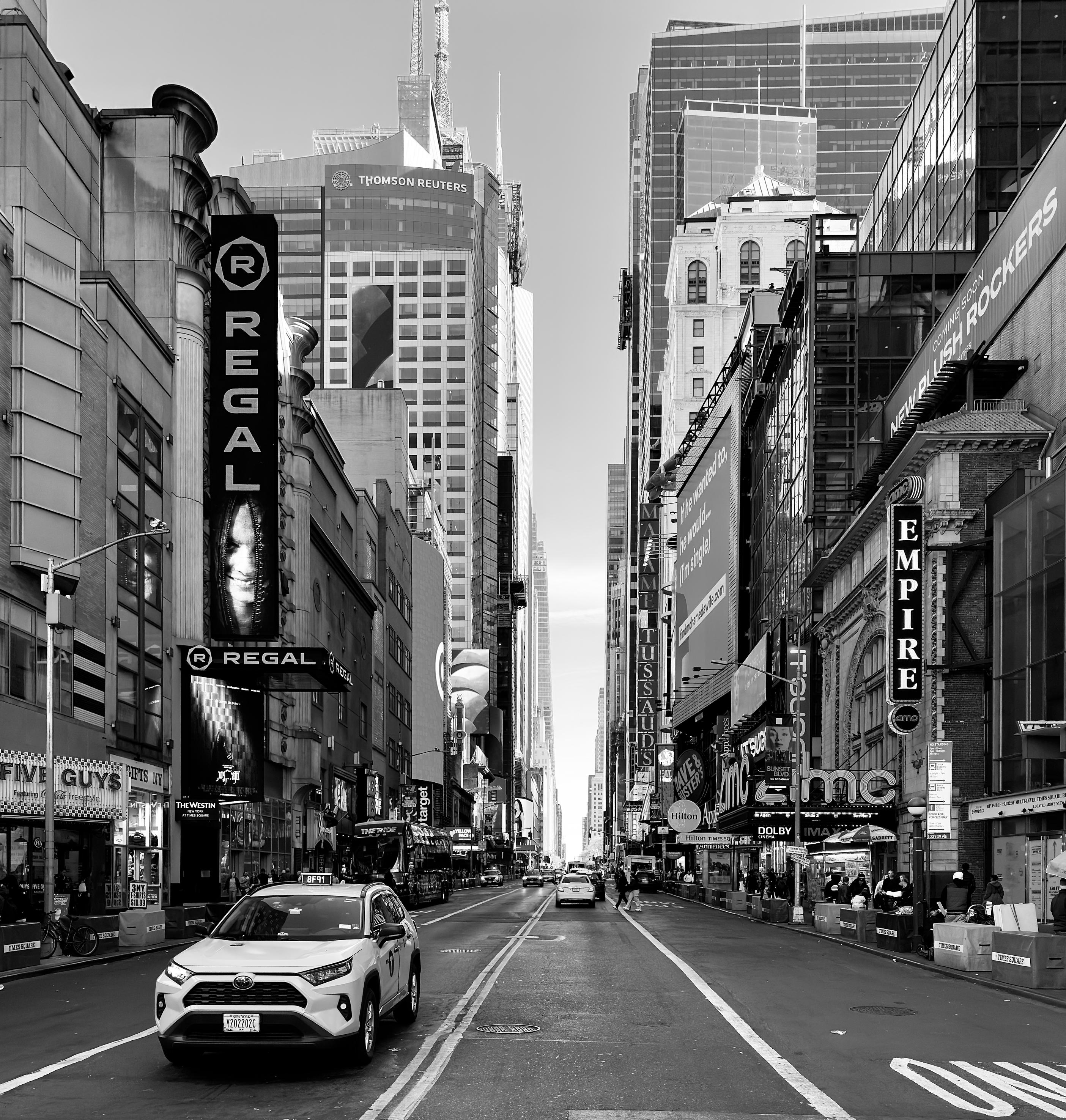A high-contrast, black and white street-level photo of West 42nd Street looking east from 8th Avenue toward Times Square in Manhattan. The street is nearly empty of traffic, with only a taxi van driving toward the camera in the left lane. A few pedestrians are visible on the sidewalks. Large, multi-story buildings with numerous electronic billboards and theater marquees line both sides of the street. On the left, Thomson Reuters tower, and a prominent Regal movie theater sign with vertical lettering and a stylized eye logo is visible. A Five Guys restaurant is also visible on the left, next to the theater. On the right, the Empire Theatre and AMC Empire 25 movie theater buildings are prominent. The overall tone is stark and geometric, emphasizing the urban canyon effect of the architecture and signage.