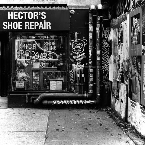 A black and white photograph in a square aspect ratio. A street-level photo out front of a narrow, urban shoe repair shop. The shop, Hector's Shoe Repair, has a large window with neon signs that read "Shoe Shine" and "Repairs." A mannequin wearing a white shirt, black hat, and glasses is visible in the window display. The building's exterior is covered in pipes, graffiti, and numerous posters. The sidewalk is cracked, and a drainpipe runs along the bottom of the wall.