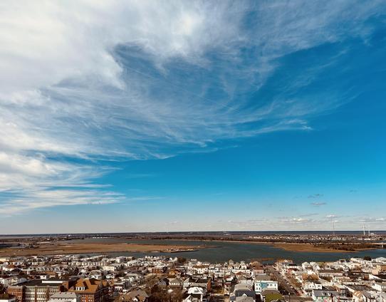 A color photograph, a high-angle landscape shot of Atlantic City facing inland under a wide, bright blue sky with wispy white clouds. The foreground shows the rooftops of the dense residential area, and beyond that is a large body of water that extends to the horizon. The water separates the town from a flat, marshy landscape in the distance. Wind turbines are visible on the horizon to the far right.