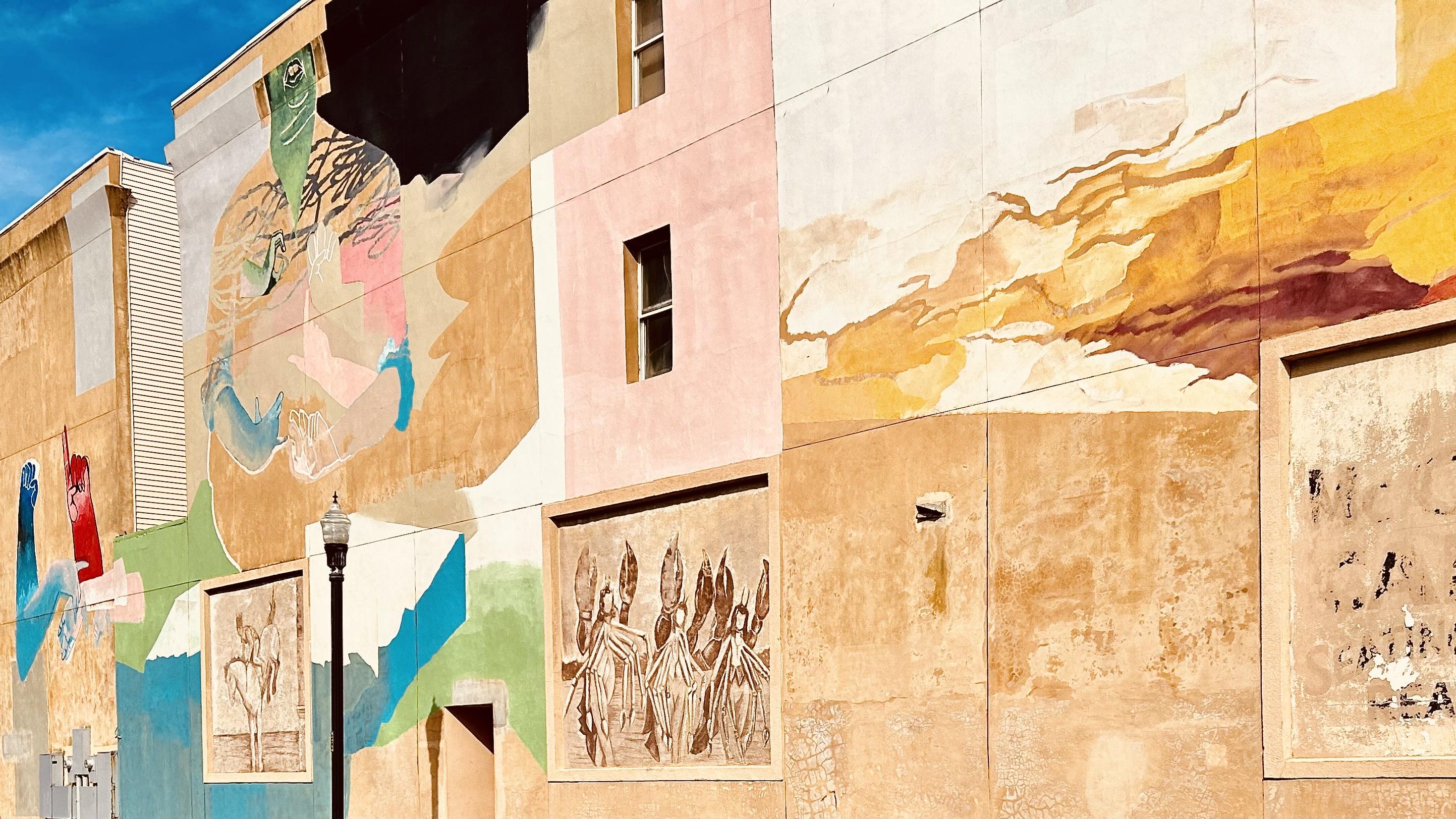 A color photograph of a large mural covering the entire side of a tan building, featuring abstract and figurative art in various colors, including blues, yellows, and reds. The mural is painted across multiple sections of the building and includes some relief elements and windows. A section of the mural near the top of the building is black and unfinished. A single lamppost is installed next to the building and the sun is casting a short shadow of the lamppost onto the building. A deep blue afternoon sky is seen above the building.