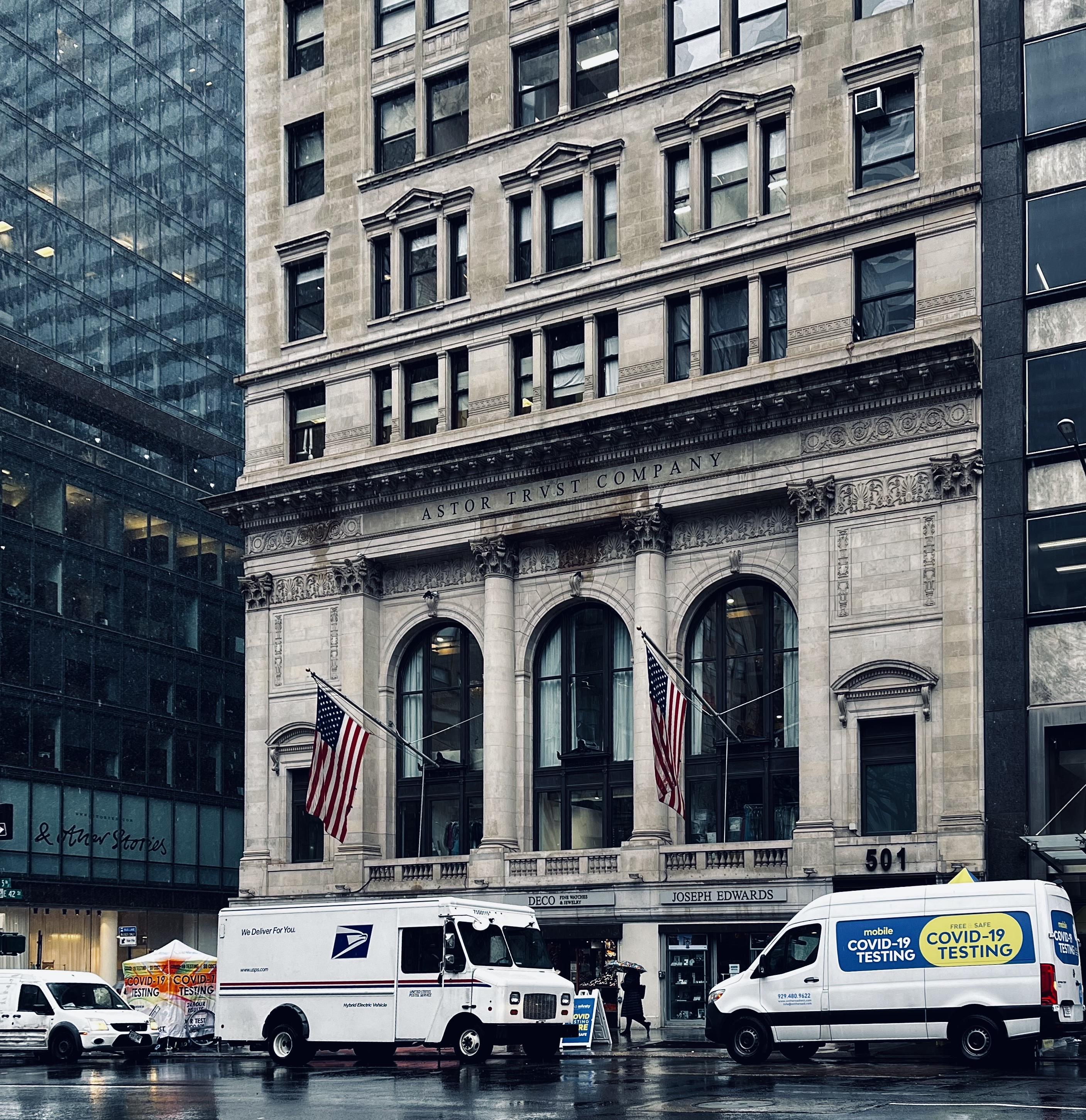A color photograph of a multi-story stone building occupying a corner lot in Midtown Manhattan on a rainy day. The rain clouds are making the scene desaturated. The building's second floor features the carved text "ASTOR TRVST COMPANY" across its facade. The street level has three large arched windows/entrances with classical columns in between, two of which display American flags. The ground floor businesses appear to be "DECO" and "JOSEPH EDWARDS." In the foreground, parked on the wet street, are two white vans: a U.S. Postal Service (USPS) delivery truck on the left, and a van on the right with prominent blue and yellow signage for "mobile COVID-19 TESTING" and "TESTING." To the far left, a white tent and a smaller white vehicle indicate a COVID-19 testing site setup on the sidewalk. Tall, modern glass buildings are on the left side of the frame.