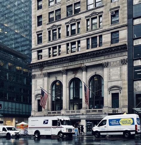 A color photograph of a multi-story stone building occupying a corner lot in Midtown Manhattan on a rainy day. The rain clouds are making the scene desaturated. The building's second floor features the carved text "ASTOR TRVST COMPANY" across its facade. The street level has three large arched windows/entrances with classical columns in between, two of which display American flags. The ground floor businesses appear to be "DECO" and "JOSEPH EDWARDS." In the foreground, parked on the wet street, are two white vans: a U.S. Postal Service (USPS) delivery truck on the left, and a van on the right with prominent blue and yellow signage for "mobile COVID-19 TESTING" and "TESTING." To the far left, a white tent and a smaller white vehicle indicate a COVID-19 testing site setup on the sidewalk. Tall, modern glass buildings are on the left side of the frame.