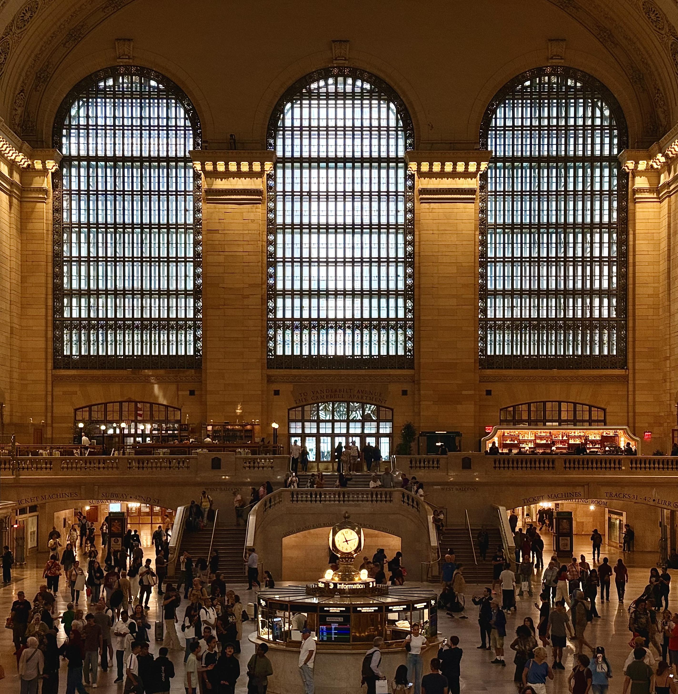 This color photograph taken inside the main concourse of Grand Central Terminal. The scene is dominated by the warm, golden-brown tones of the stone walls and the dramatic overcast morning light streaming into the space through three massive, arched windows that span the upper section of the image. The windows are composed of many small, gridded panes, which strongly filter the natural light. The main level is a large, open area with many people. In the center foreground, there is a circular information booth, topped with a four-sided brass clock. Behind the information booth, a grand staircase splits and ascends to an upper level, where balconies overlook the concourse. More people are visible on these staircases and balconies.
