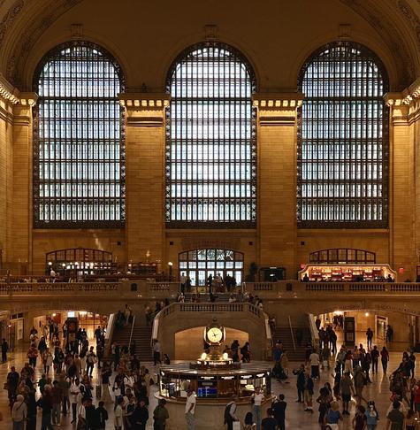 This color photograph taken inside the main concourse of Grand Central Terminal. The scene is dominated by the warm, golden-brown tones of the stone walls and the dramatic overcast morning light streaming into the space through three massive, arched windows that span the upper section of the image. The windows are composed of many small, gridded panes, which strongly filter the natural light. The main level is a large, open area with many people. In the center foreground, there is a circular information booth, topped with a four-sided brass clock. Behind the information booth, a grand staircase splits and ascends to an upper level, where balconies overlook the concourse. More people are visible on these staircases and balconies.