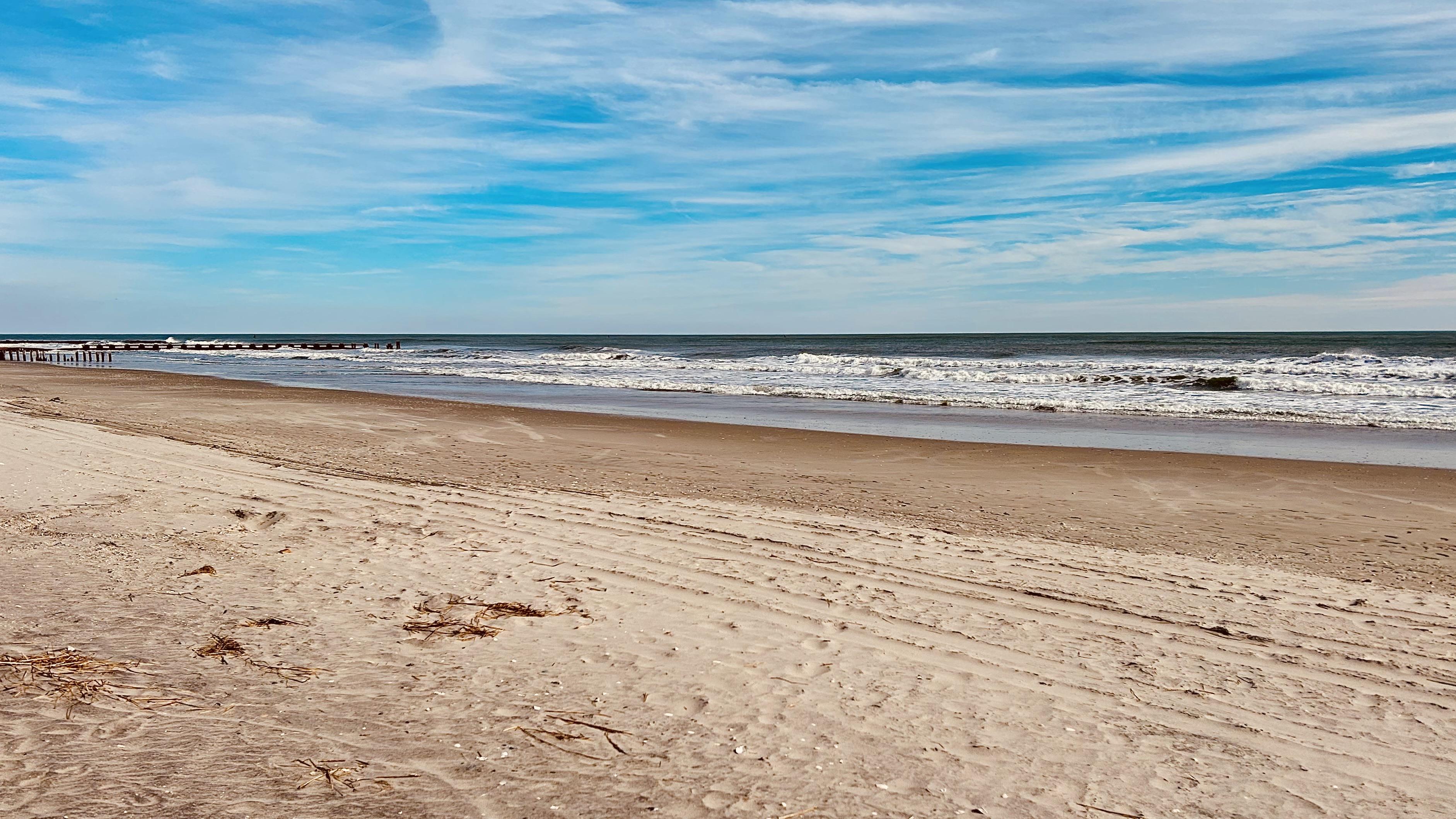 A color photograph, a panoramic view of an Atlantic City beach and the Atlantic Ocean under a partially cloudy, bright blue sky. The foreground consists of wide, light tan sand showing subtle tire marks from an ATV. Moving toward the water, the sand transitions to a darker, damp band at the tide line. The ocean surface is a deep slate blue, meeting the beach with white, churning foam from moderate waves. The sky is a vibrant blue with streaks of wispy white cirrus and darker, more defined cumulus clouds.