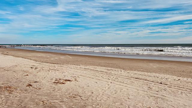 A color photograph, a panoramic view of an Atlantic City beach and the Atlantic Ocean under a partially cloudy, bright blue sky. The foreground consists of wide, light tan sand showing subtle tire marks from an ATV. Moving toward the water, the sand transitions to a darker, damp band at the tide line. The ocean surface is a deep slate blue, meeting the beach with white, churning foam from moderate waves. The sky is a vibrant blue with streaks of wispy white cirrus and darker, more defined cumulus clouds.