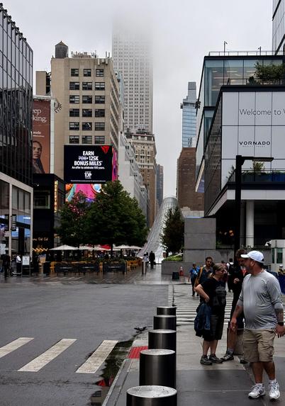A color photograph of the overcast and foggy street scene in midtown Manhattan. The view looks down a street lined with modern and older high-rise buildings. In the center, the top floors of the Empire State Building are partially obscured by low-hanging fog. A large digital billboard on the left side of the street displays an advertisement with red and white text. Below the billboard, there is an outdoor dining area with yellow umbrellas and several patrons. On the right, a modern glass building has a sign that reads "Welcome to VORNADO." In the immediate foreground, a group of three people is walking on the sidewalk; one man in a white hat and grey shirt is closest to the camera. The street is wet from rain, and there are several silver bollards lining the edge of the sidewalk and a crosswalk marked on the street. The overall mood is cool and slightly muted due to the weather.
