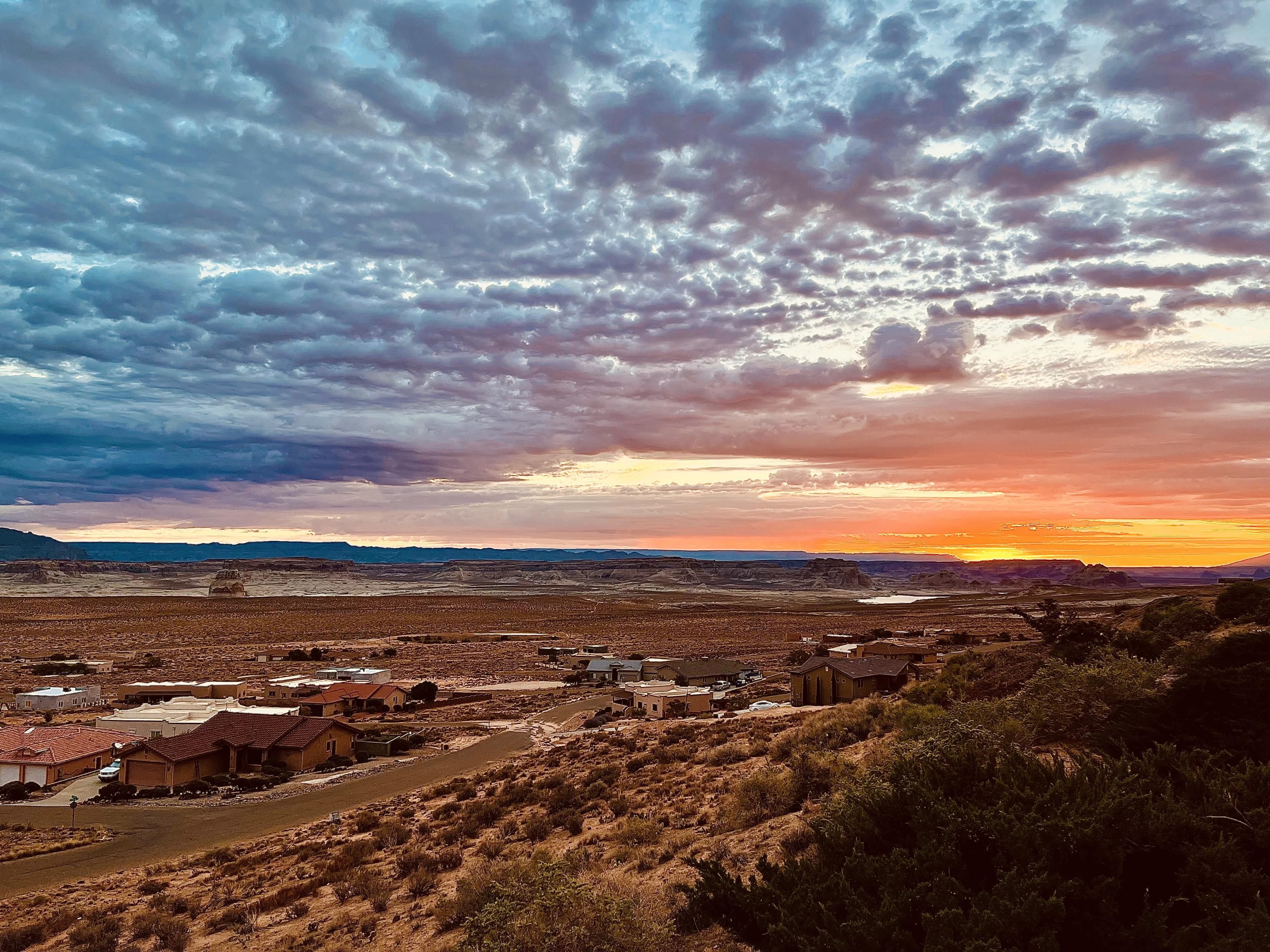 A color photography, a wide-angle landscape shot of a dramatic sunrise over a small desert community in Greenehaven, Arizona. The foreground is dominated by dry, brush-covered hills sloping down toward a cluster of modern, earth-toned stucco houses. Beyond the houses, the flat, arid landscape stretches toward a distant mesa. The sky is the main focus, with an expansive, heavily textured layer of altocumulus and stratus clouds. The clouds on the left are a deep, moody blue and gray, while the right side of the sky is vibrant with the warm glow of the rising sun, featuring intense oranges, reds, and soft yellows just above the horizon. The light casts a warm, soft glow over the landscape.