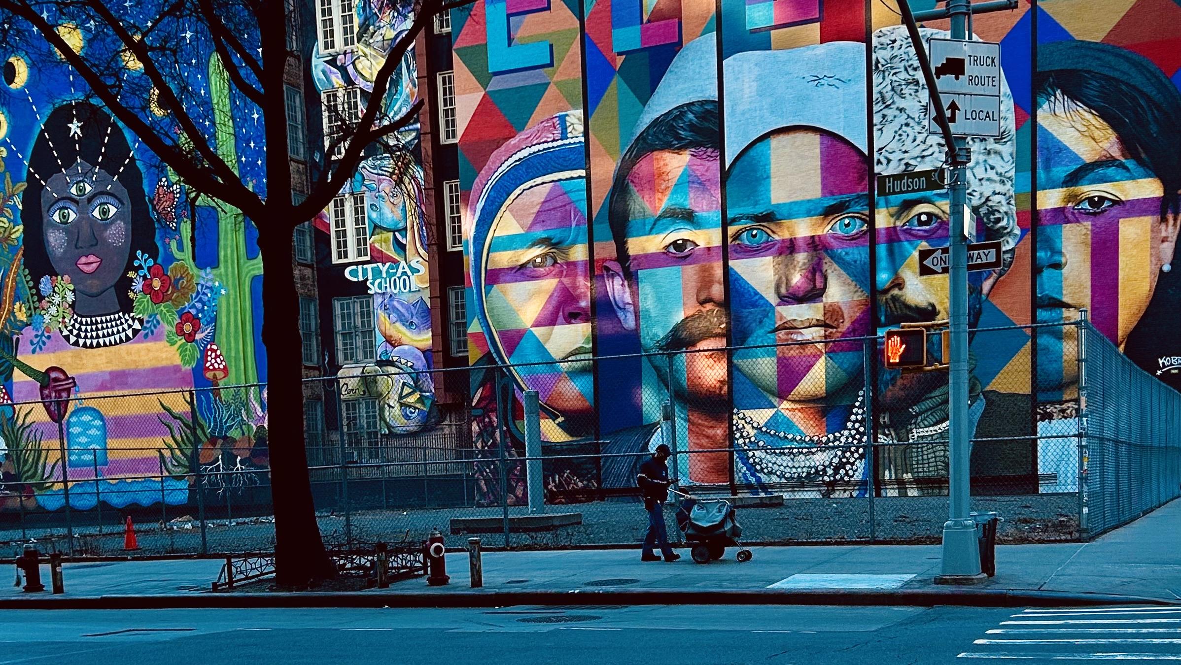 A color, wide-angle photograph of a school that sits on a corner lot in the West Village neighborhood of Manhattan. An elaborate mural on the side of the building dominates the scene, featuring a sequence of large-scale portraits. Between the building and the camera is a playground enclosed with a chain-link fence. The mural is painted in a vibrant, geometric style with strong colors like blue, red, and yellow, creating an abstract, mosaic-like effect on the faces. On the far left is a separate, more figurative mural of a woman with a dark complexion, decorated with flowers, stars, and a third eye on her forehead. In the foreground, a postal delivery person walks on the sidewalk, pushing a mail cart. A small sign on a pole near the center of the image indicates a Truck Route and a street sign for Hudson St and One Way. The street and foreground shadows are bathed in a deep, cool blue light, contrasting with the warm, vibrant colors of the artwork above.