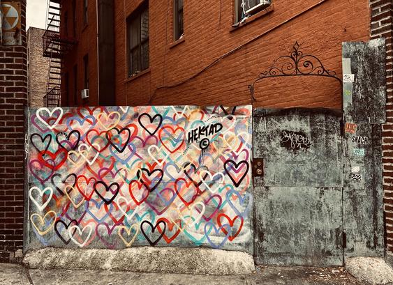 A color photograph taken along Broadway in the Inwood neighborhood of Manhattan on an overcast winter day. The focal point of the scene is a vibrant mural of intertwined hearts painted on a low, corrugated metal wall, located in a narrow vacant lot between two brick buildings. The hearts are outlined in various colors, including red, white, black, purple, teal, and yellow, layered over each other to create a dense, colorful pattern. Near the top center of the mural, the tag "HEKTAD" is visible in black. To the right is a reddish brown brick apartment building with a black metal fire escape visible. To the left is a brown brick building with a fallout shelter sign. To the right of the mural is a tall, distressed metal gate painted a dull gray-green. This gate has various black and white graffiti tags on it, and its top edge features an ornate, wrought-iron scrollwork decoration.