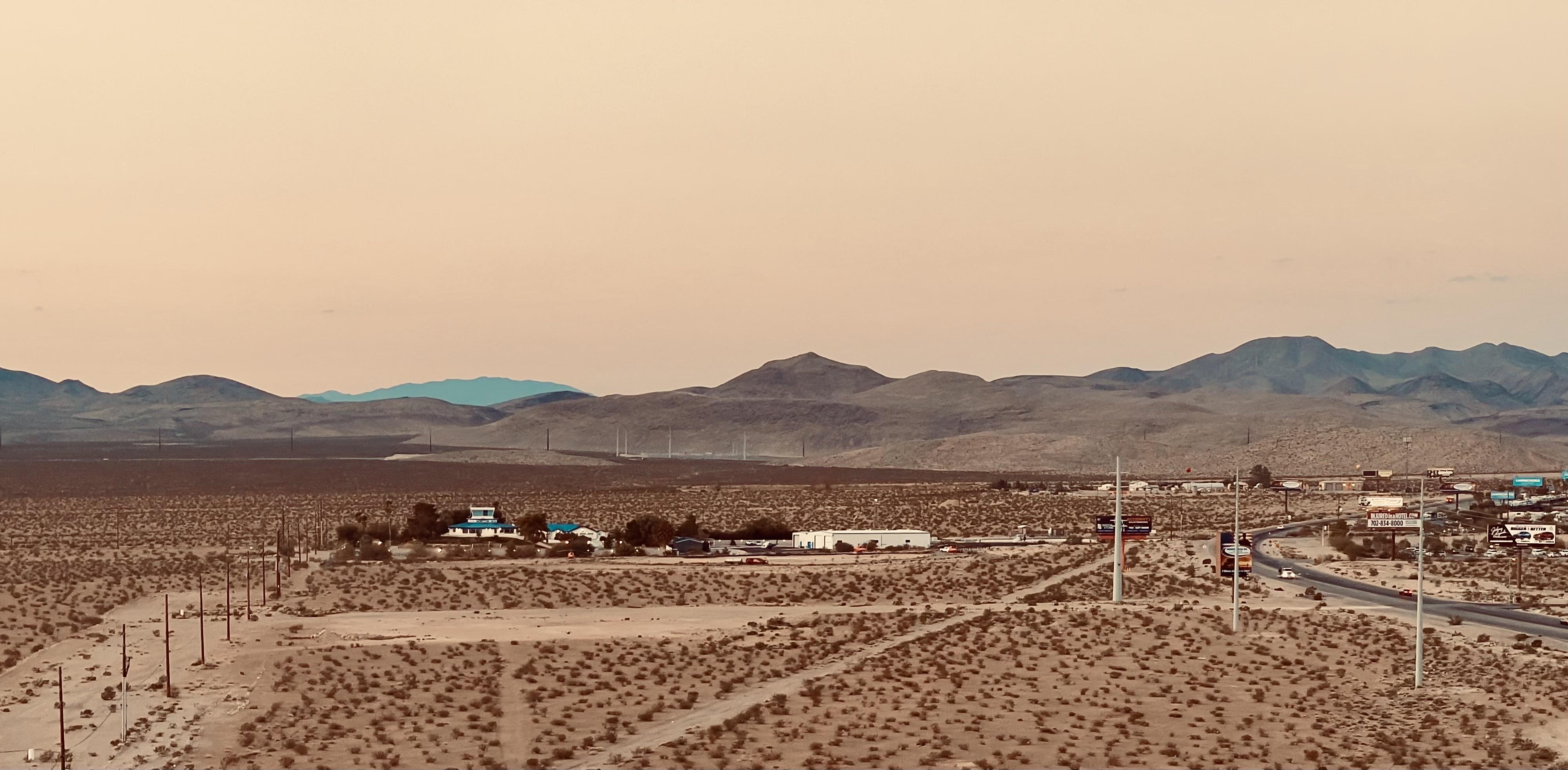 A color, landscape photograph taken at sunrise on the outskirts of Las Vegas. The scene is dominated by warm, sepia-toned light and rolling desert mountains in the background. The foreground features patchy scrub, utility poles, and the curve of a multi-lane highway. A small, bright blue-and-white roadside outpost is visible amid a scattering of low-lying commercial buildings and signs near the road. The atmosphere is hazy and muted.