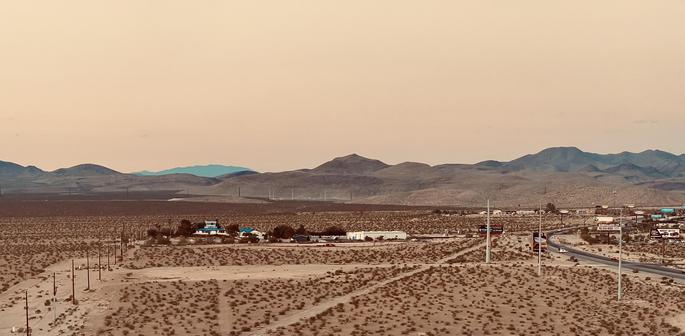 A color, landscape photograph taken at sunrise on the outskirts of Las Vegas. The scene is dominated by warm, sepia-toned light and rolling desert mountains in the background. The foreground features patchy scrub, utility poles, and the curve of a multi-lane highway. A small, bright blue-and-white roadside outpost is visible amid a scattering of low-lying commercial buildings and signs near the road. The atmosphere is hazy and muted.