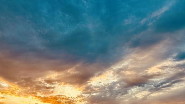 A color photograph of a vibrant, dramatic sky at sunset above Tuba City, Navajo Nation. The composition is split diagonally by color. The upper two-thirds of the image is dominated by a deep, rich cyan/teal blue with scattered, darker clouds. The lower third features brilliant, warm tones of orange, gold, and deep violet, which indicates the location of the setting sun just below the lower left corner of the frame. Rays of bright, golden light are visible streaking up from the bottom left corner through the warmer clouds, which transition into the cooler, darker colors above. The overall impression is one of intense color contrast and dynamic atmosphere.