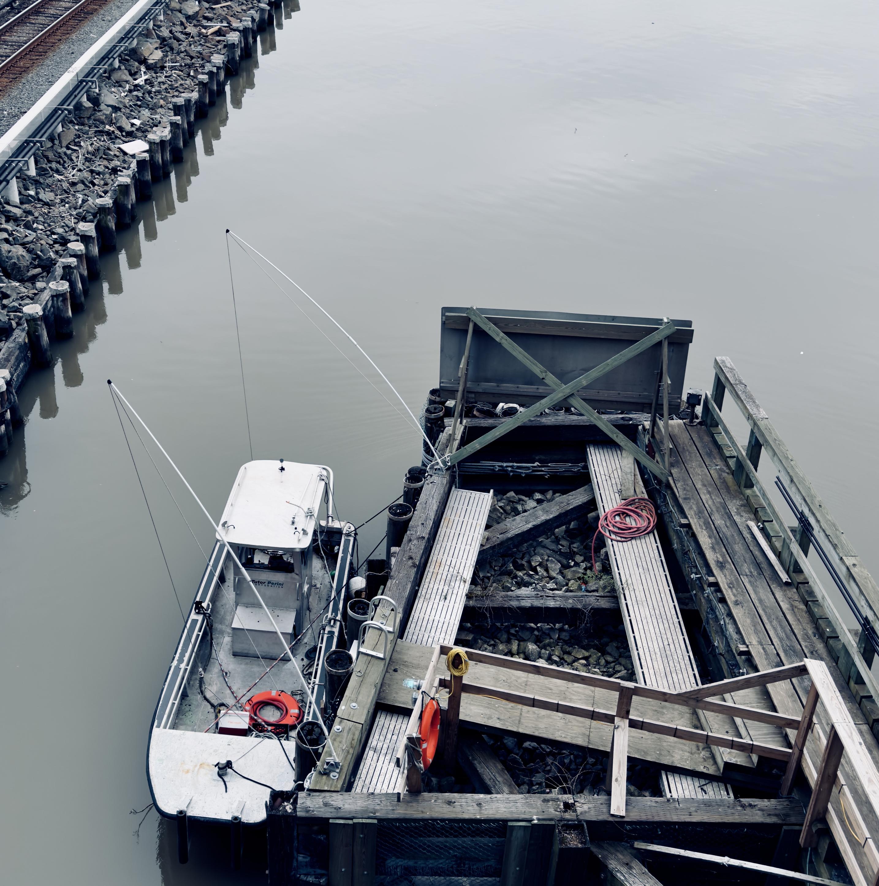 A color photograph in a square aspect ratio taken on an overcast morning. An overhead view of a small white utility boat docked at a wooden pier attached to the University Heights Bridge on the grey, calm water of the Harlem River. The boat, positioned on the left, has an orange life preserver visible near the stern. The pier on the right is made of weathered wood planks and beams, with loose rocks filling part of the dock's surface between two wooden ramps. A coiled red hose and another orange life preserver are visible on the pier. To the far left, a retaining wall of wooden pilings runs parallel to the river, topped by a stretch of rocky, dark riprap and a set of railway tracks.