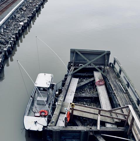 A color photograph in a square aspect ratio taken on an overcast morning. An overhead view of a small white utility boat docked at a wooden pier attached to the University Heights Bridge on the grey, calm water of the Harlem River. The boat, positioned on the left, has an orange life preserver visible near the stern. The pier on the right is made of weathered wood planks and beams, with loose rocks filling part of the dock's surface between two wooden ramps. A coiled red hose and another orange life preserver are visible on the pier. To the far left, a retaining wall of wooden pilings runs parallel to the river, topped by a stretch of rocky, dark riprap and a set of railway tracks.