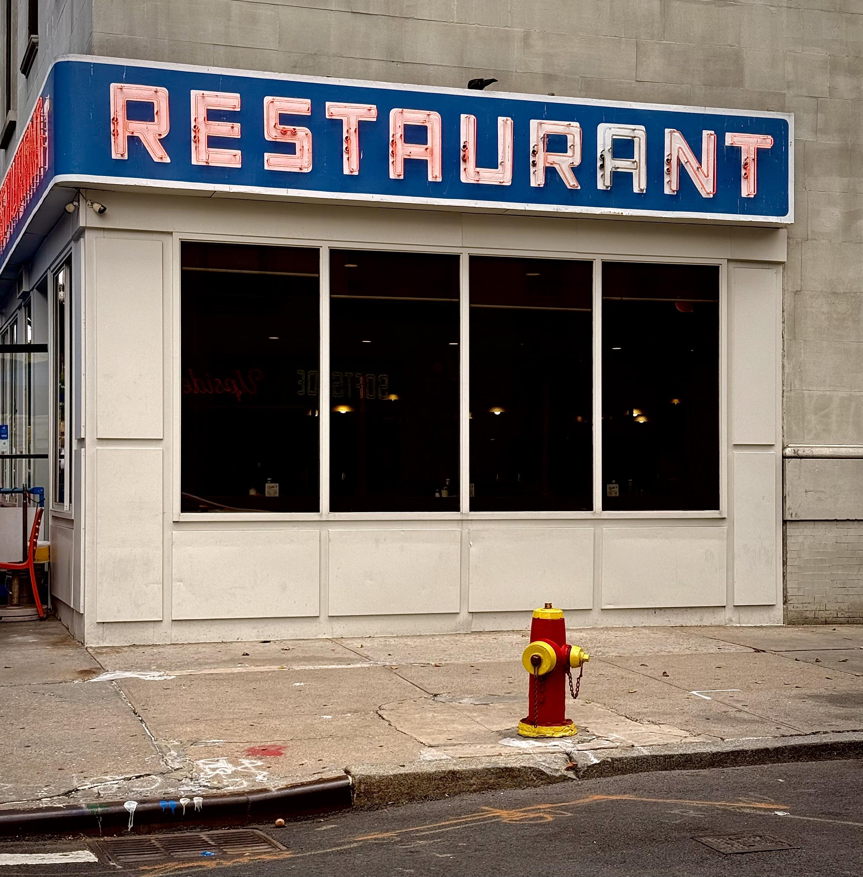 A color photograph in a square aspect ratio. The focal point of the scene is the facade of a classic diner-style restaurant building. Dominating the upper section is a large, rectangular, neon sign with a bright blue background panel and white trim. The word "RESTAURANT" is displayed prominently in a block, sans-serif font using neon tubing. The "R," "E," "S," and "A" are illuminated in a pink or reddish-pink neon, contrasting with the remaining "T," "U," "R," "A," "N," and "T," which appear unlit (or dimly lit) white or light pink in the center. A large, horizontal window bank, composed of four equally-sized, dark-tinted rectangular panes occupies the center. The darkness of the glass makes the interior appear nearly black, with only a few indistinct, warm-toned lights visible far back. Faint, backwards-reflected text from a sign across the street can be seen in the glass. The restaurant sits directly on a typical New York City sidewalk made of broken, uneven grey concrete slabs. A vibrant, short yellow fire hydrant with a red cap and chain is positioned near the curb, slightly off-center to the right of the building's front.