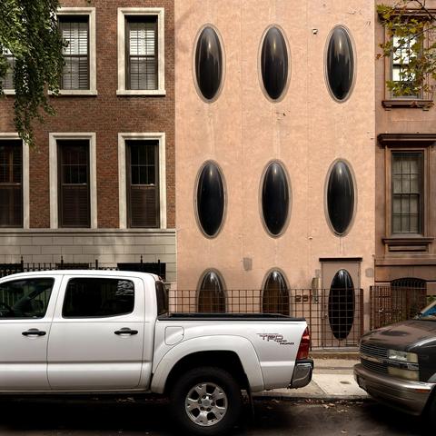 A color photograph in a square aspect ratio, taken along East 71st Street on an overcast morning. The focal point of the scene is a row of three homes. On the left and right sides of frame are traditional dark colored brownstones. In the center is a lighter tan colored building with three floors of vertically oriented, tinted, oval windows that immediately stand out. On the street in front of the homes is a silver pickup truck and a dark colored van parked.