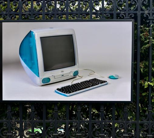 A color photograph in a square aspect ratio. Taken along Fifth Avenue outside of the Cooper Hewitt design art museum on an overcast day. The focal point of the image is a large poster size image of a bondi blue G3 iMac, keyboard, and mouse on a white background. The poster has a black frame and is attached to a black wrought iron fence. Behind the fence many green trees fill the frame.