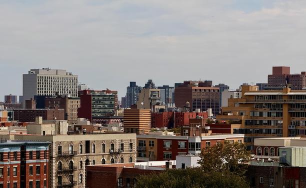 A color photograph, a panoramic view of the dense Harlem cityscape taken from an elevated observation deck in Morningside Park on a cloudy day. The scene is dominated by an array of buildings ranging in height and architectural style. The foreground consists of the rooftops of shorter, older residential and commercial buildings, many have fire escapes on their facades. Notable colors in the foreground are the dark red and brown bricks of these structures, with one building on the far left having a distinctive blue and red lower facade. Taller trees from the park are seen on the lower right corner of the frame.