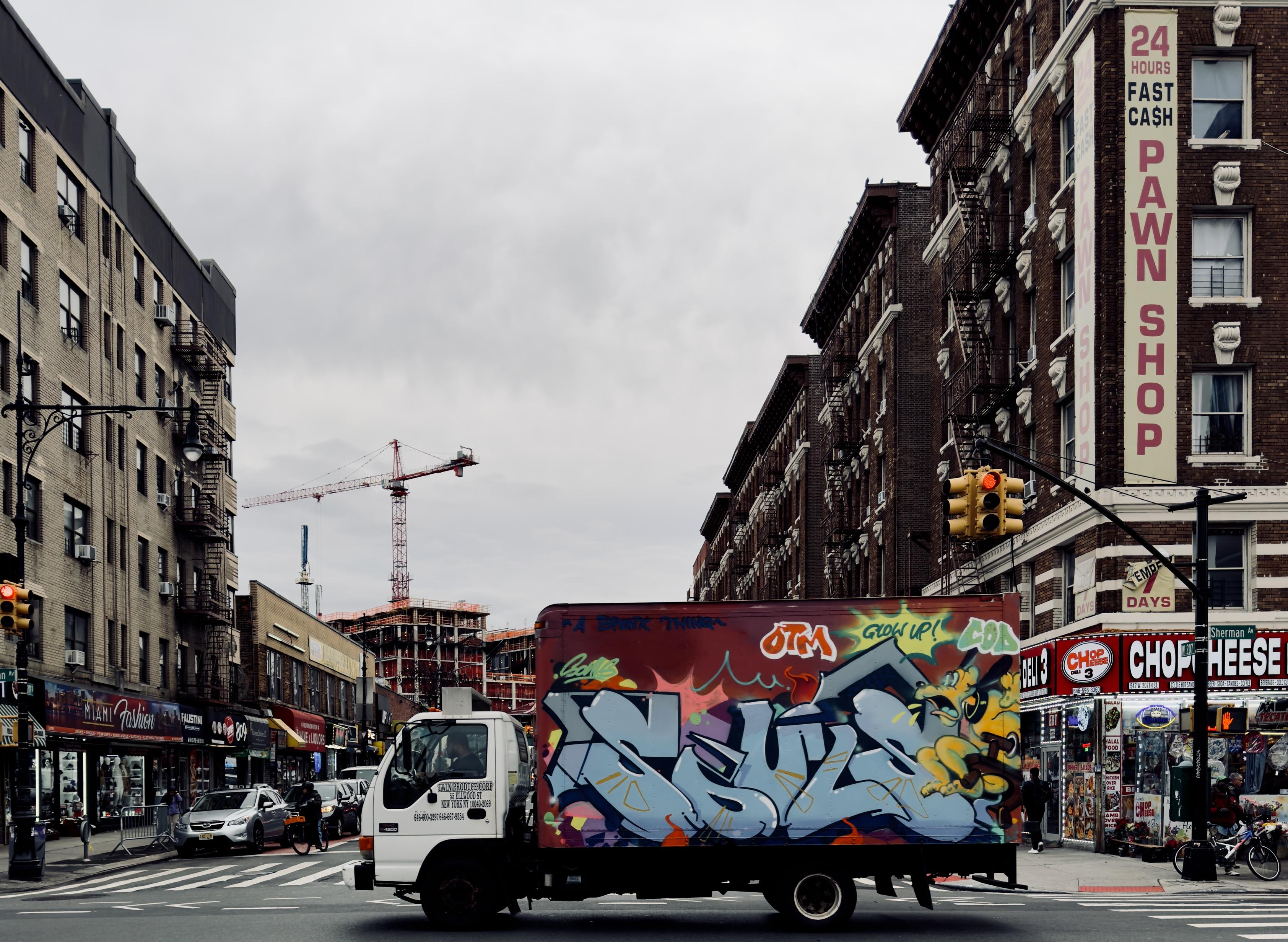 A color photograph taken on a cloudy day in Inwood, Manhattan. Centered on the intersection of 207th Street and Sherman Ave. The central focus is a delivery truck moving left-to-right across the foreground, with its entire cargo box covered in vibrant graffiti. The graffiti features large, stylized letters in light blue and white, outlined in black, with a yellow cartoon-like character on the right side. To the left and right, the street is lined with multi-story, reddish-brown brick apartment buildings. The buildings on the right feature ground-floor storefronts, including a prominent sign for a "24 Hours fast cash pawn shop" that runs vertically up the building's facade. Traffic lights are visible overhead to the right, showing red. In the mid-ground, a construction site is visible down the street. A tall red and white construction crane dominates the skyline in this middle section. The sky is overcast and gray. Cars are parked or moving on the street in the background, and various smaller signs and storefronts are visible on both sides.