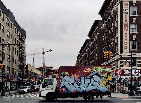 A color photograph taken on a cloudy day in Inwood, Manhattan. Centered on the intersection of 207th Street and Sherman Ave. The central focus is a delivery truck moving left-to-right across the foreground, with its entire cargo box covered in vibrant graffiti. The graffiti features large, stylized letters in light blue and white, outlined in black, with a yellow cartoon-like character on the right side. To the left and right, the street is lined with multi-story, reddish-brown brick apartment buildings. The buildings on the right feature ground-floor storefronts, including a prominent sign for a "24 Hours fast cash pawn shop" that runs vertically up the building's facade. Traffic lights are visible overhead to the right, showing red. In the mid-ground, a construction site is visible down the street. A tall red and white construction crane dominates the skyline in this middle section. The sky is overcast and gray. Cars are parked or moving on the street in the background, and various smaller signs and storefronts are visible on both sides.