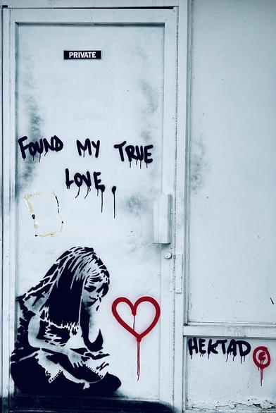 A color photograph in portrait orientation of a black-and-white stencil-style artwork on a distressed white exterior door of an office building. The door also has the word "PRIVATE" in white text on a small black sign near the top. The artwork features a seated young woman in a dress, looking down with her chin rested on her palm near her face. To her right is a simple red outline of a heart, with a drip of red paint trailing down from the point. Above the woman and the heart are the words "FOUND MY TRUE LOVE," painted in a dripping, black, graffiti-style script. Below the words are a few small black drips. On the white wall to the right of the door, near the bottom, are the capitalized, dark red words "HEKTAD" and a small red circle with an 'C' inside it to symbolize copyright. It is late afternoon on a cloudy day so the temperature of the image is cool.