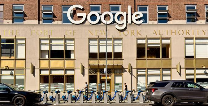 A color photograph of the Google office building located in the Chelsea neighborhood of Manhattan, New York City. The building is a large, former transportation office with a brick facade on the upper level and a light-colored stone lower level. Prominently displayed above the second-story windows are large, white, dimensional letters spelling "Google." Beneath the "Google" sign, across the light-colored facade, is gold-colored lettering that reads "THE PORT OF NEW YORK AUTHORITY," indicating the building's original, historical use. The lower level features several tall, rectangular windows with gold-colored frames. A central entryway is recessed, flanked by more windows. In front of the building, a row of blue-and-white Citi Bike bicycles are docked, running almost the entire width of the frame. Two pedestrians are visible near the building entrance and bikes. On the street in front of the bike rack, a black sedan is parked on the left, and a gray SUV is parked on the right. The upper portion of the building shows a row of brick with several open, angled windows. It is noon on a clear day so sharp shadows appear in the scene.